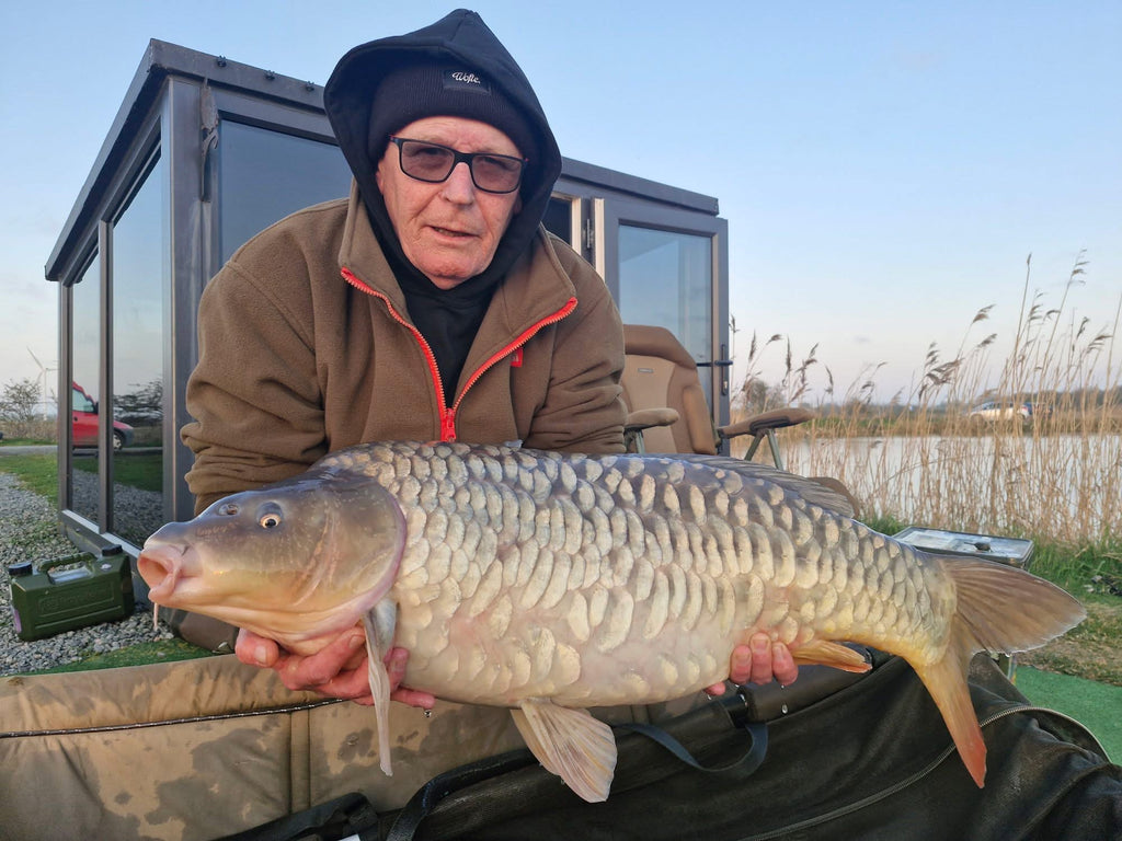 Man holding a large fish by a lake with a building and shed in the background