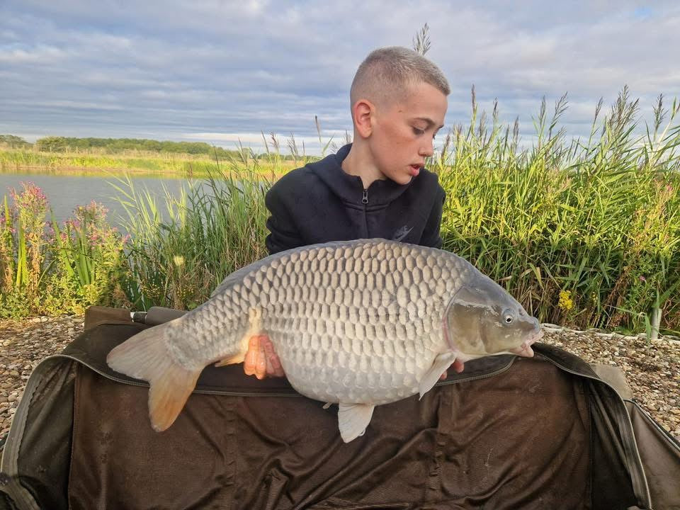 Person holding a large fish by a lake with grass and sky in the background