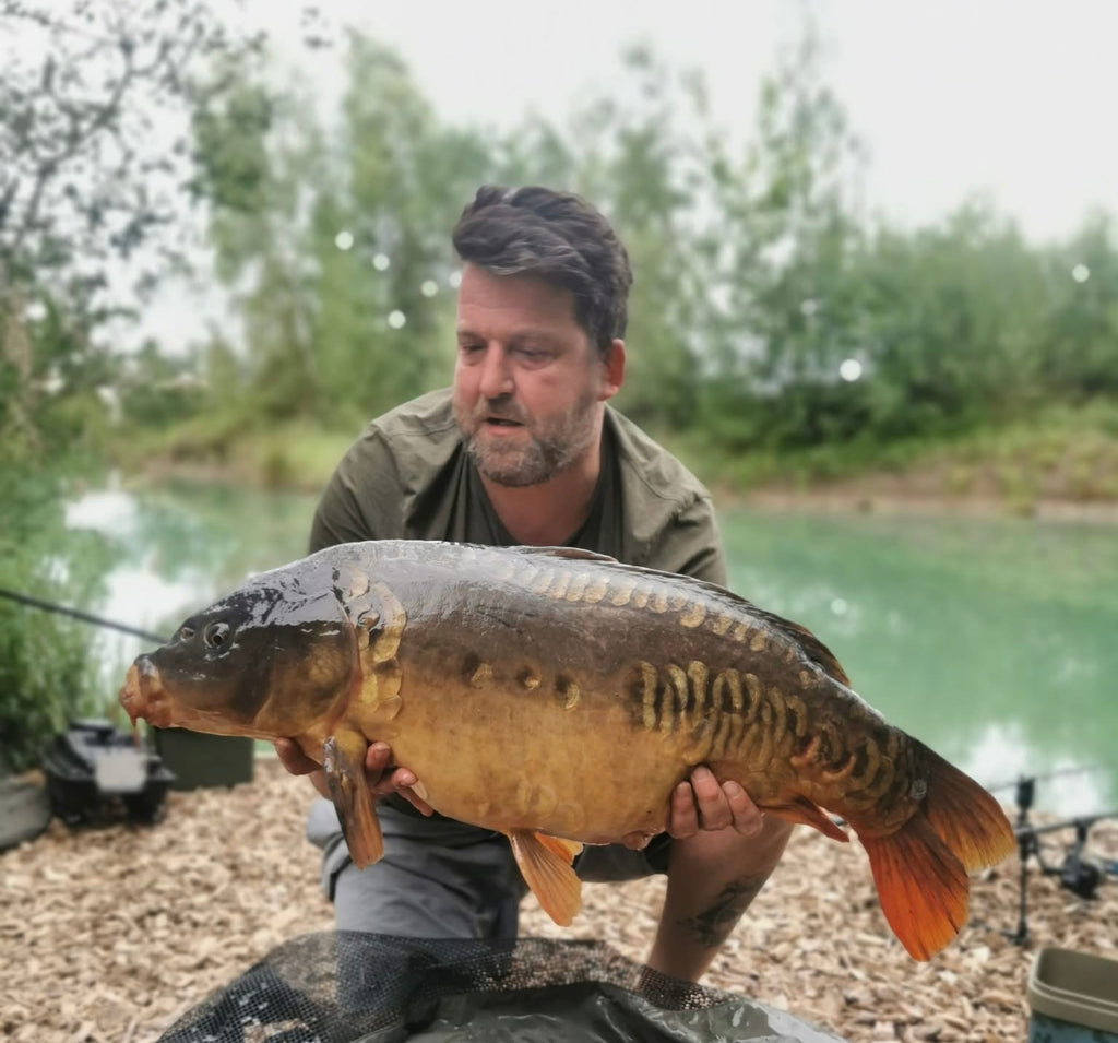Man holding a large fish by a lake with trees in the background