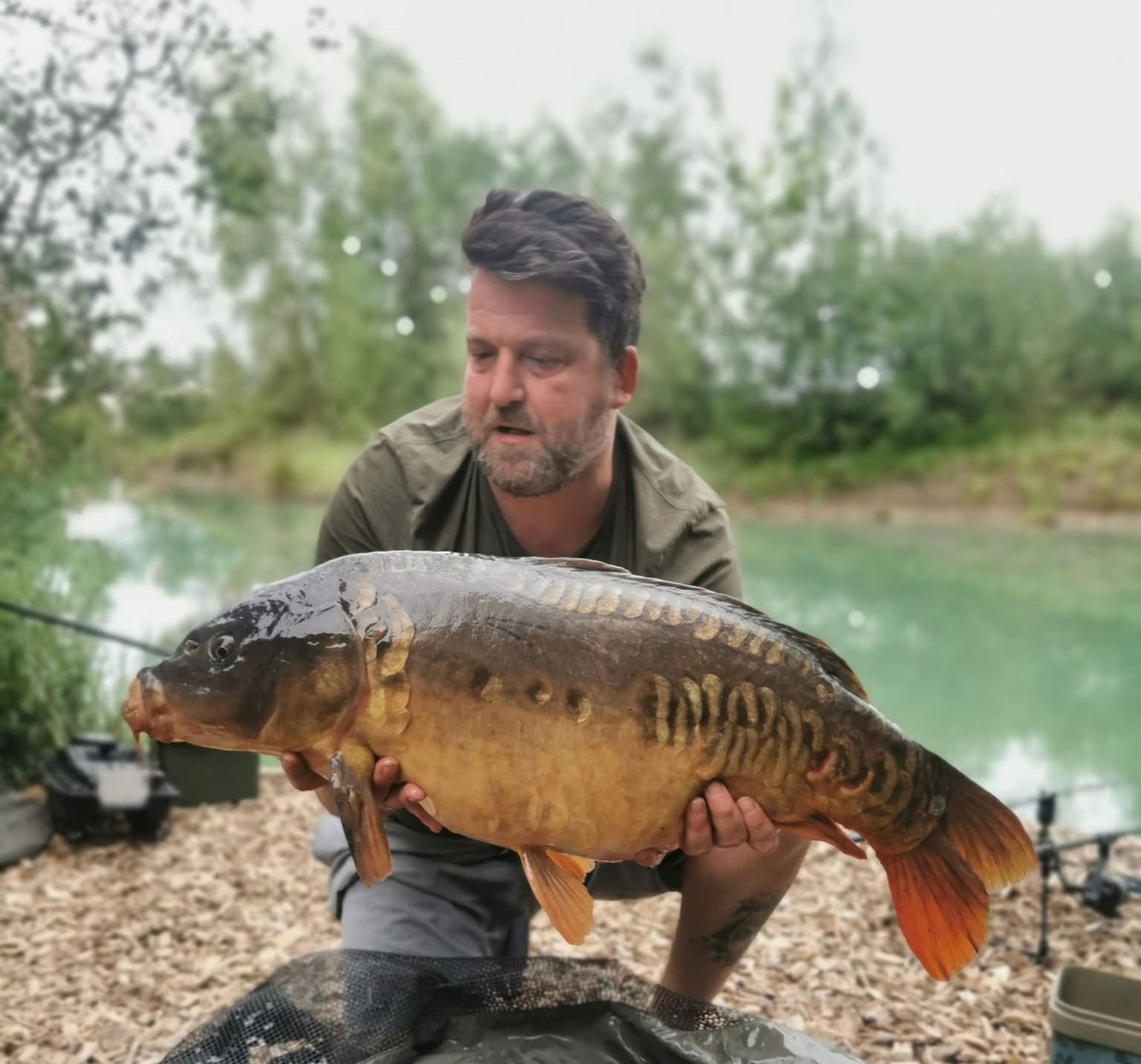 Man holding a large fish by a lake with trees in the background